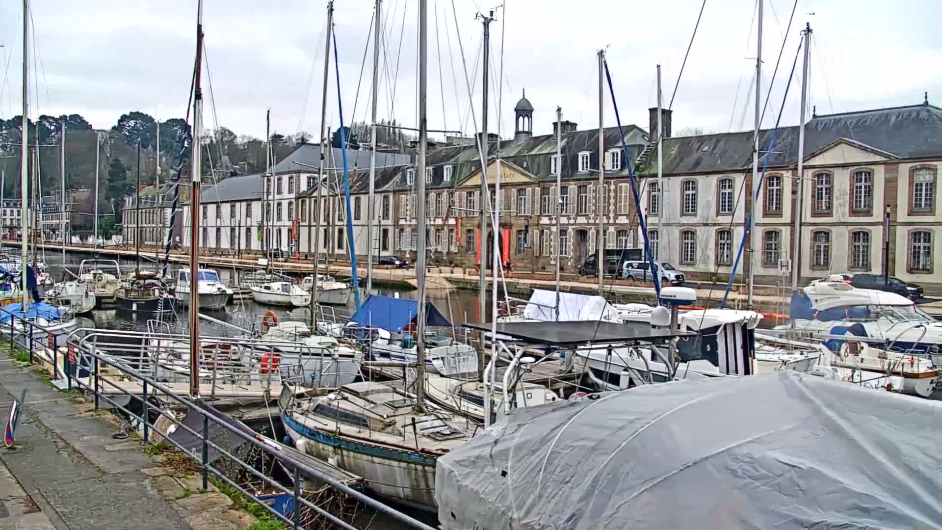 An overcast day view from a boat's rigging overlooks a marina with several boats docked, traditional buildings lining the waterfront, and a wooded hill in the background.