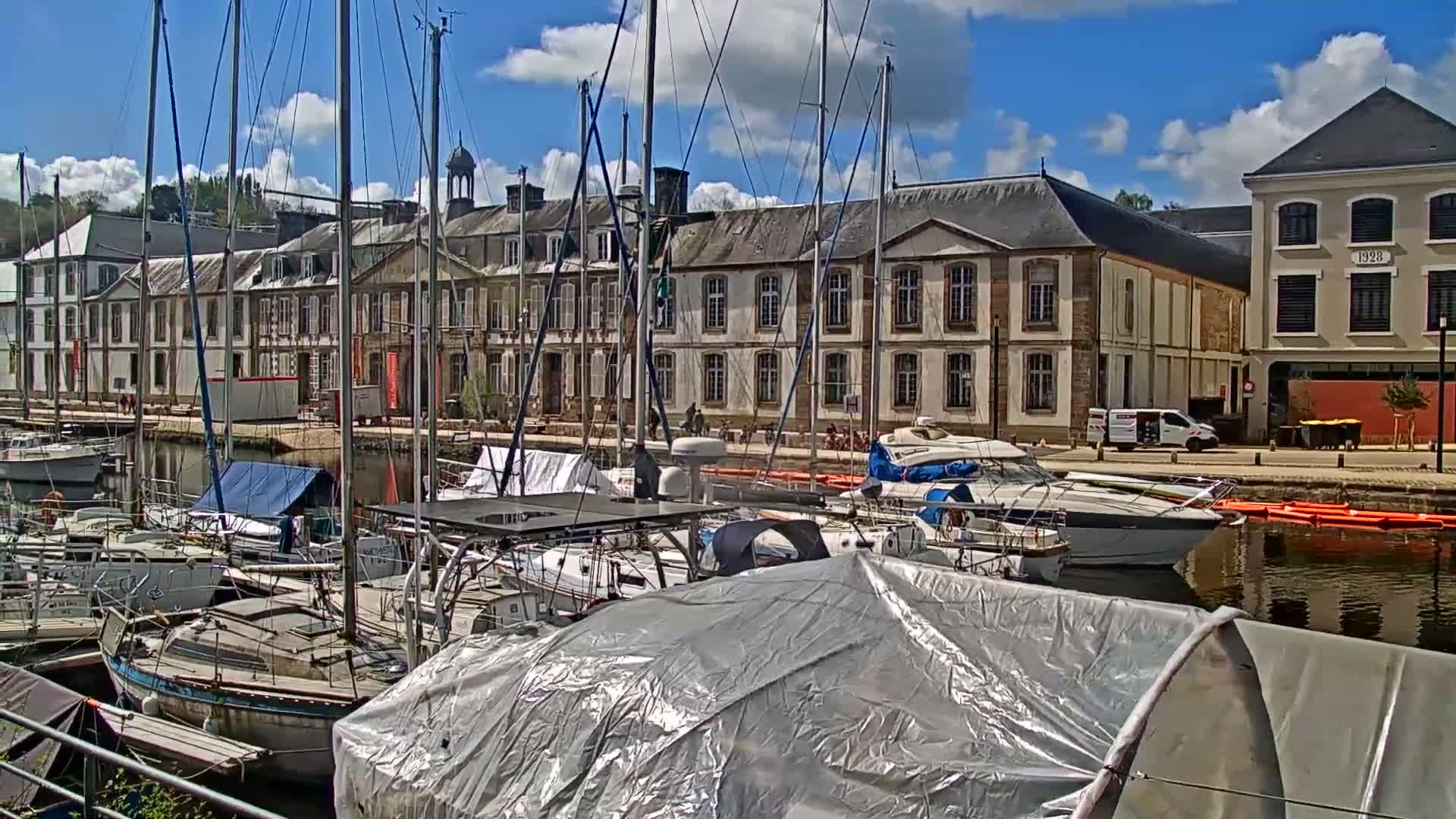An overcast day view from a boat's rigging overlooks a marina with several boats docked, traditional buildings lining the waterfront, and a wooded hill in the background.