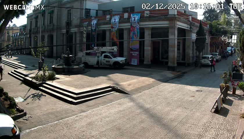 A bright, sunny day illuminates an urban street scene featuring historical-looking buildings with columns, a prominent fountain, steps, a white utility truck, various parked cars, and pedestrians walking on the paved surfaces.