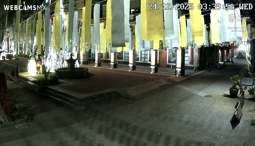 An outdoor scene at night shows a large, light-colored tent dominating the foreground, with an illuminated building featuring columns and murals, and a dimly lit street visible in the background under clear skies.