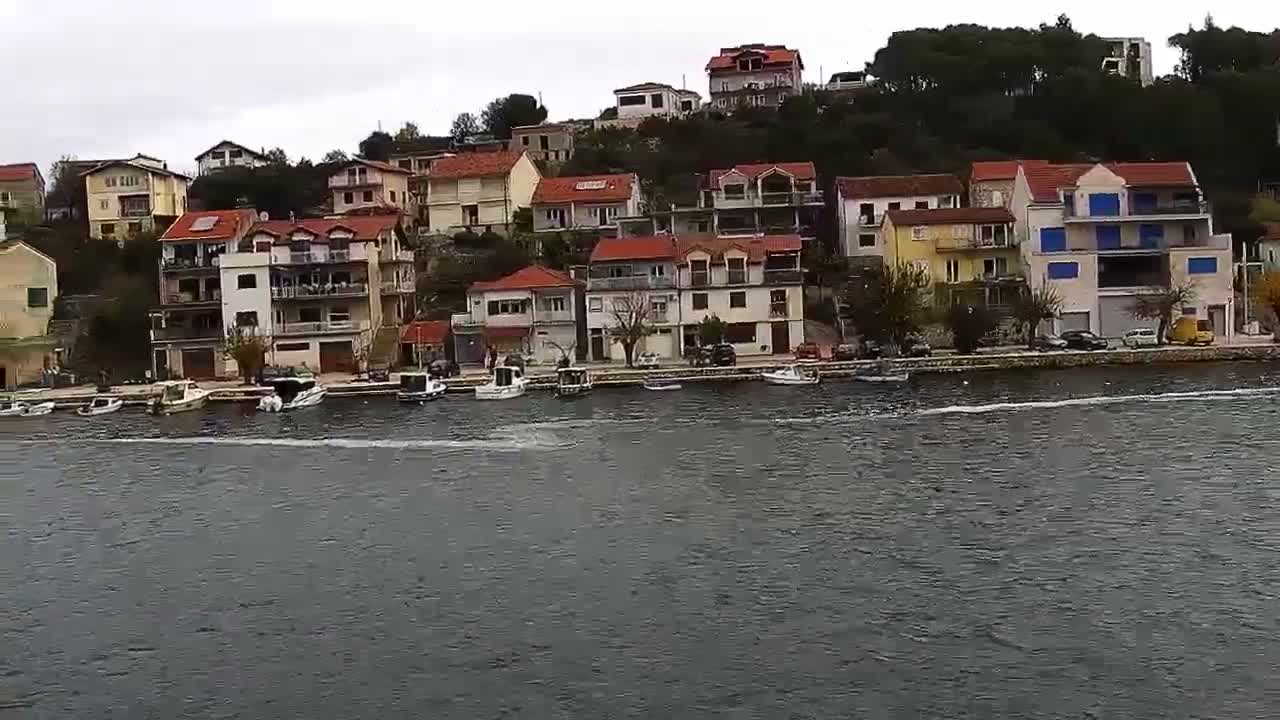 Under a grey, overcast sky, a coastal town with numerous multi-story houses and red-tiled roofs cascades down a hillside to a waterfront lined with docked boats, all set against dark, choppy water.