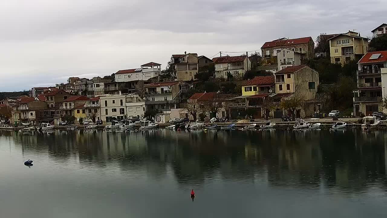 Under a grey, overcast sky, a coastal town with numerous multi-story houses and red-tiled roofs cascades down a hillside to a waterfront lined with docked boats, all set against dark, choppy water.