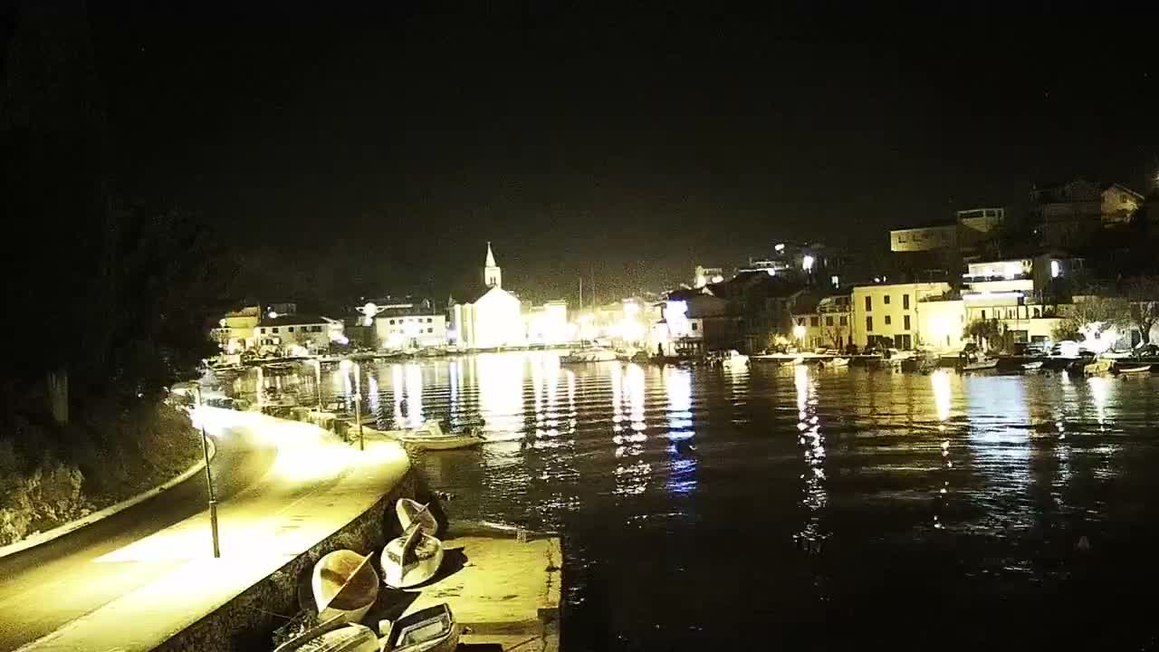 Under a grey, overcast sky, a coastal town with numerous multi-story houses and red-tiled roofs cascades down a hillside to a waterfront lined with docked boats, all set against dark, choppy water.