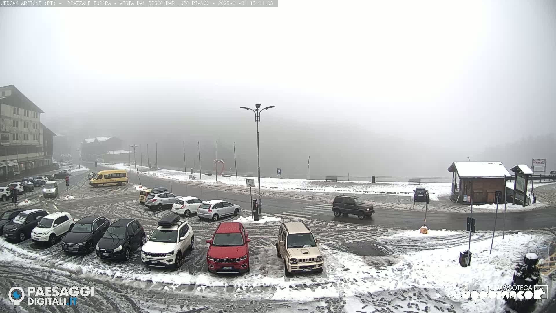 A snow-covered parking lot with several cars and a bus, situated in a foggy, snowy environment.