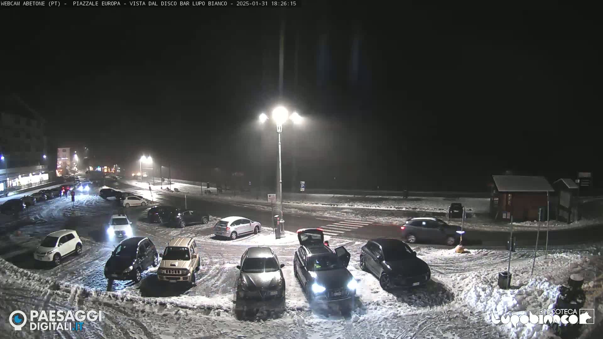 A snow-covered parking lot at night with several cars parked, illuminated by streetlights.