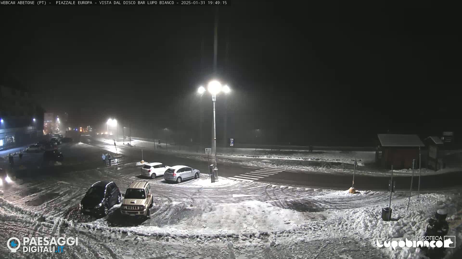 A nighttime snow scene shows a mostly empty parking lot and roadway with several cars parked, under lightly falling snow.