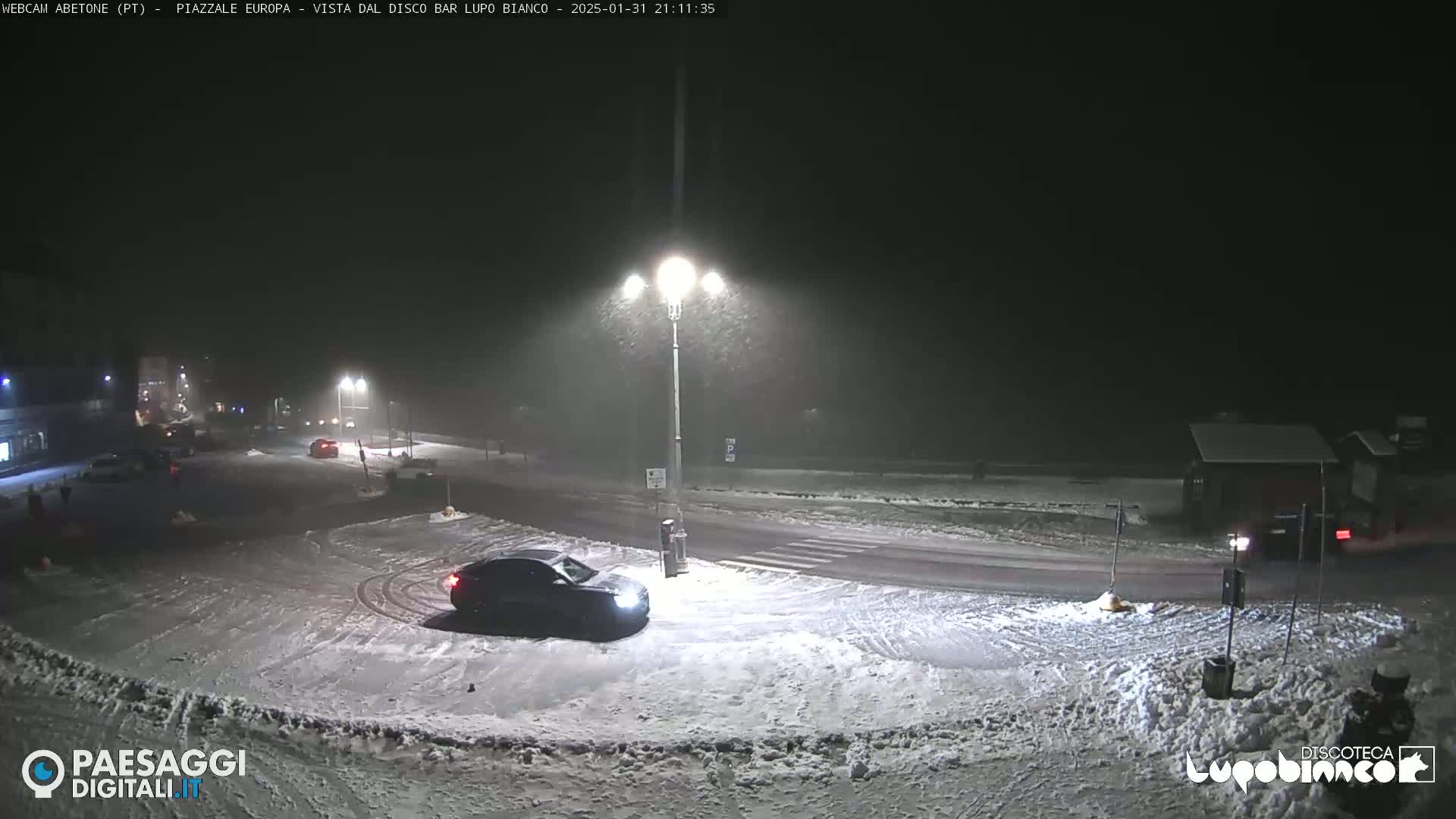 A snow-covered roadway at night with a car parked near a streetlight, and several other vehicles visible in the distance under falling snow.