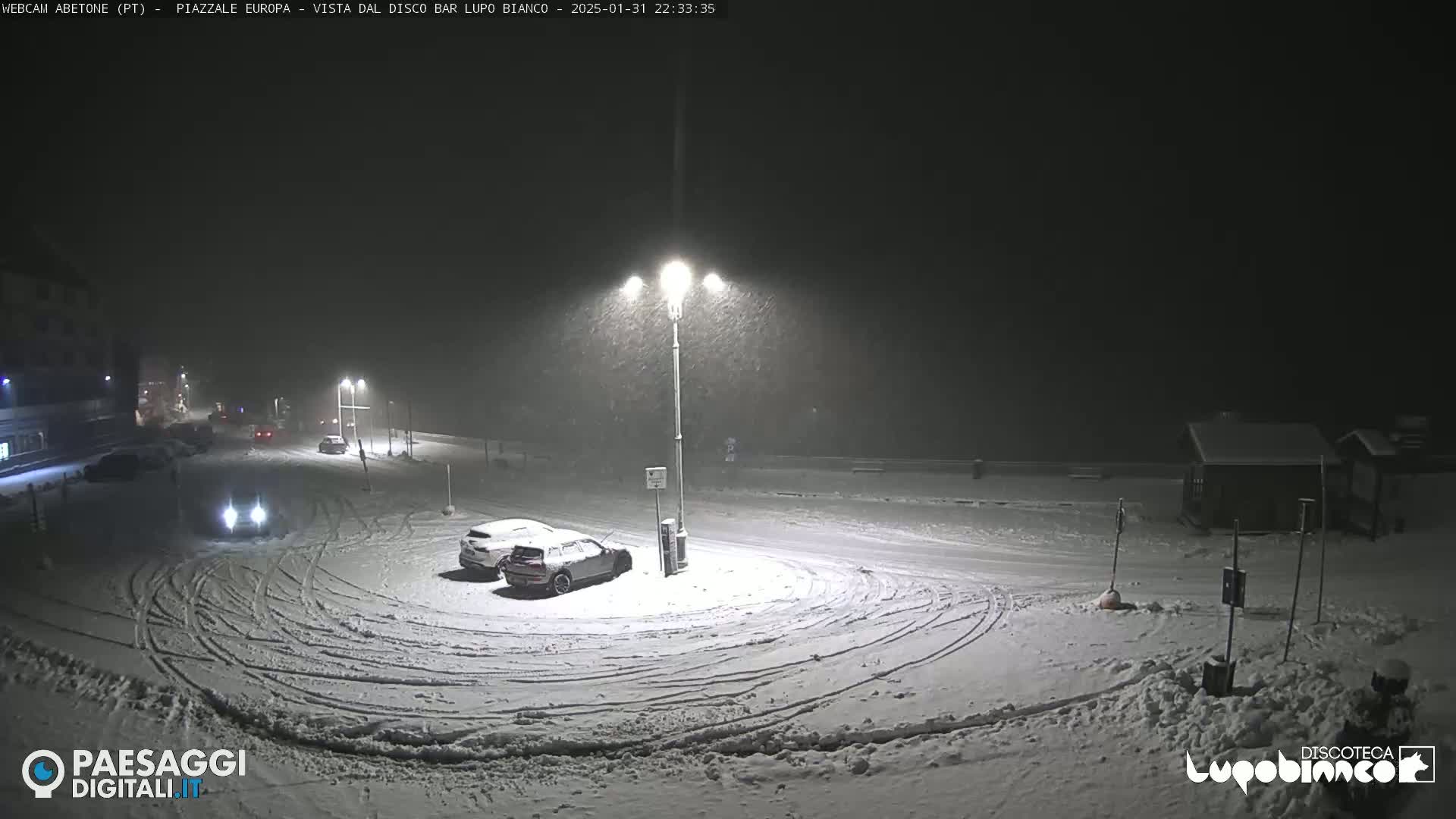 A snow-covered parking lot with several cars is illuminated by streetlights during a nighttime snowstorm.