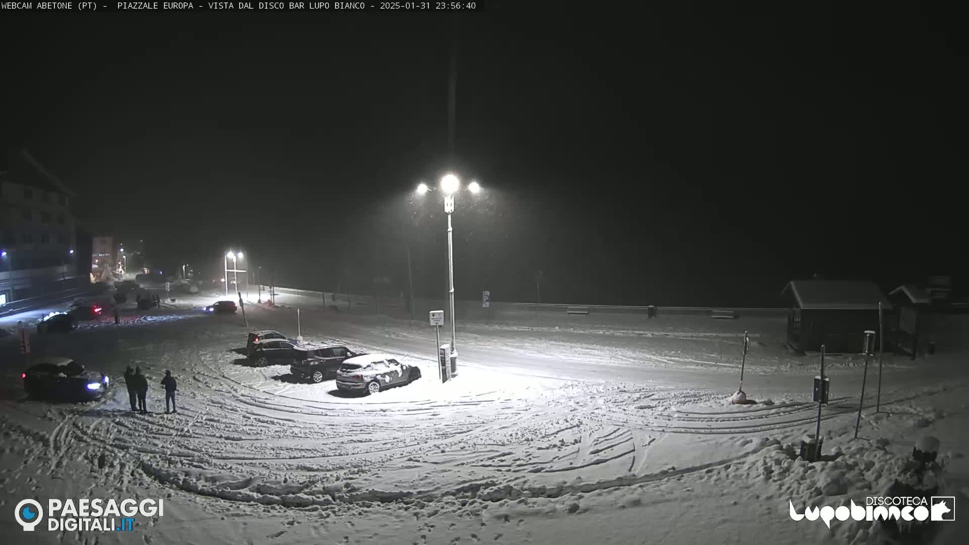 A snow-covered parking lot at night with several cars parked and a few people standing near them under lightly falling snow.