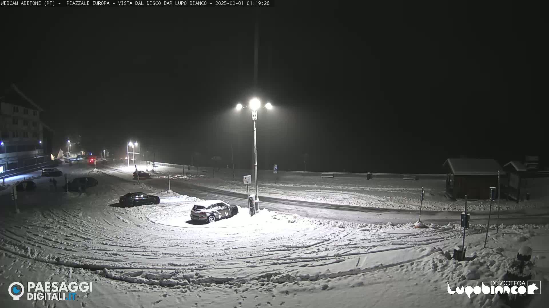 A snow-covered parking lot and roadway are illuminated at night by streetlights under a dark, snowy sky.