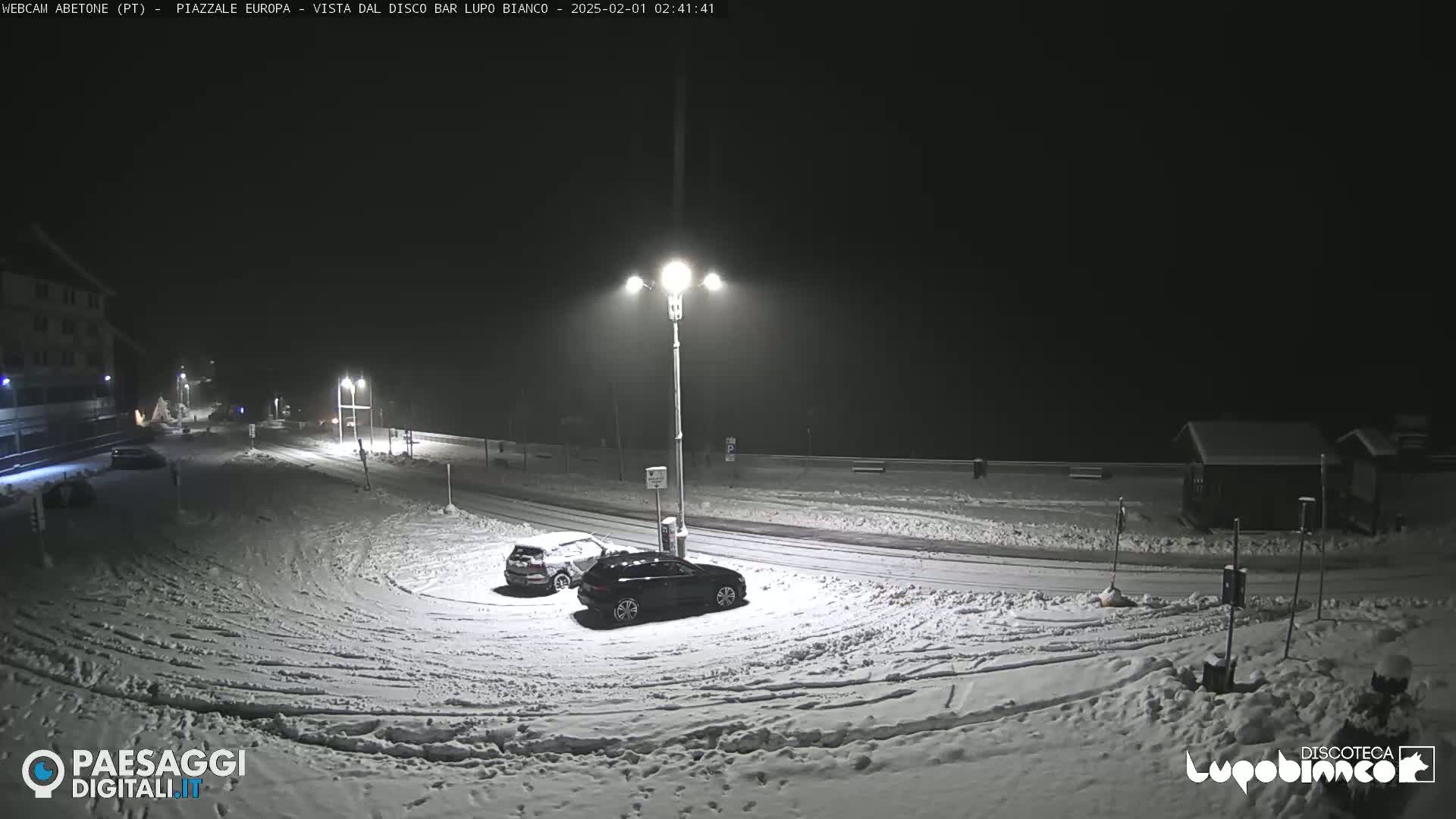 A snow-covered parking lot at night, illuminated by streetlights, with two cars parked and a building in the background.