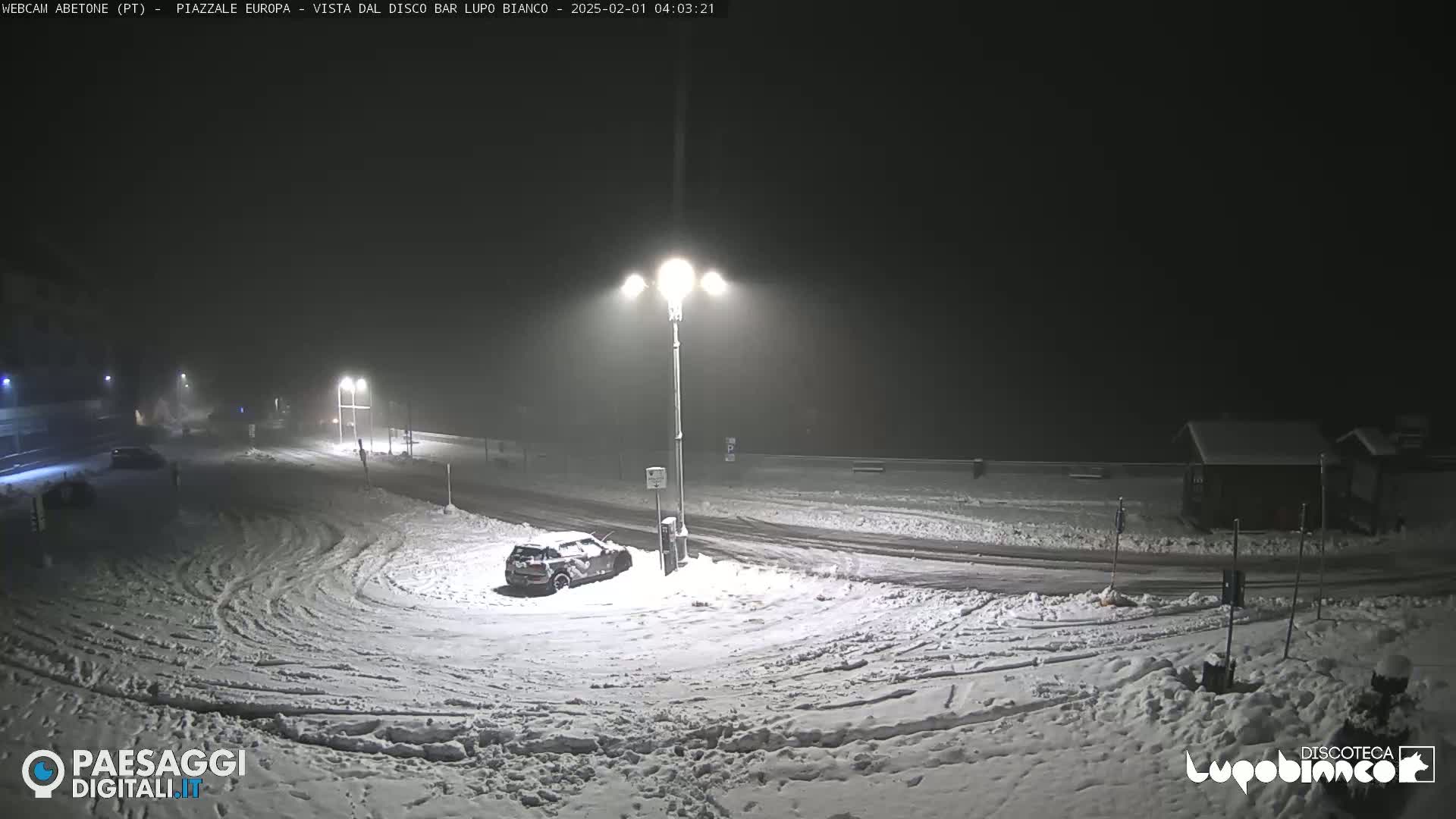 A snow-covered parking lot and road are illuminated by streetlights at night in foggy conditions, with a few vehicles visible.