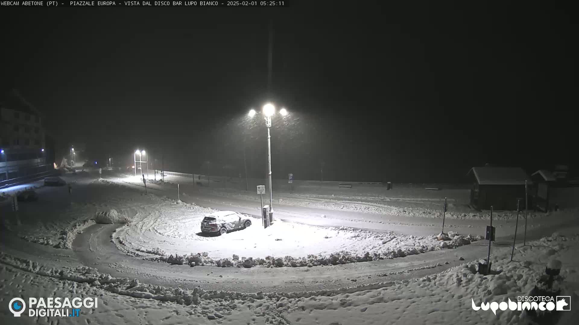 A snow-covered street scene at night, illuminated by streetlights, with a car parked in the snow and falling snow.