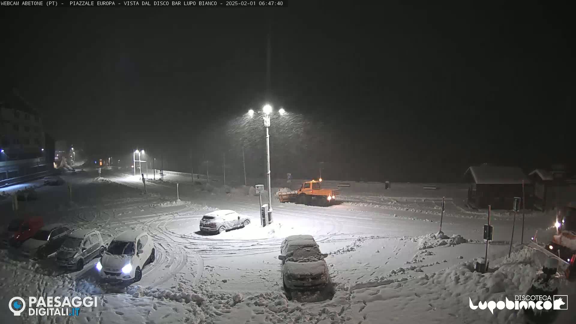 A snow-covered parking lot at night is being plowed by a small snowplow during a heavy snowfall.