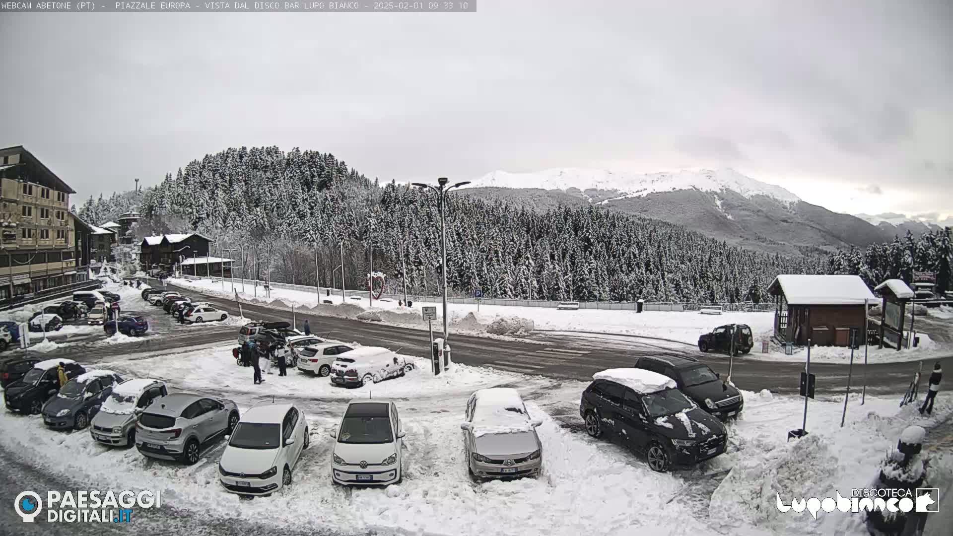 A snow-covered parking lot with cars and a few people, situated in front of a snow-covered forest and mountains under an overcast sky.