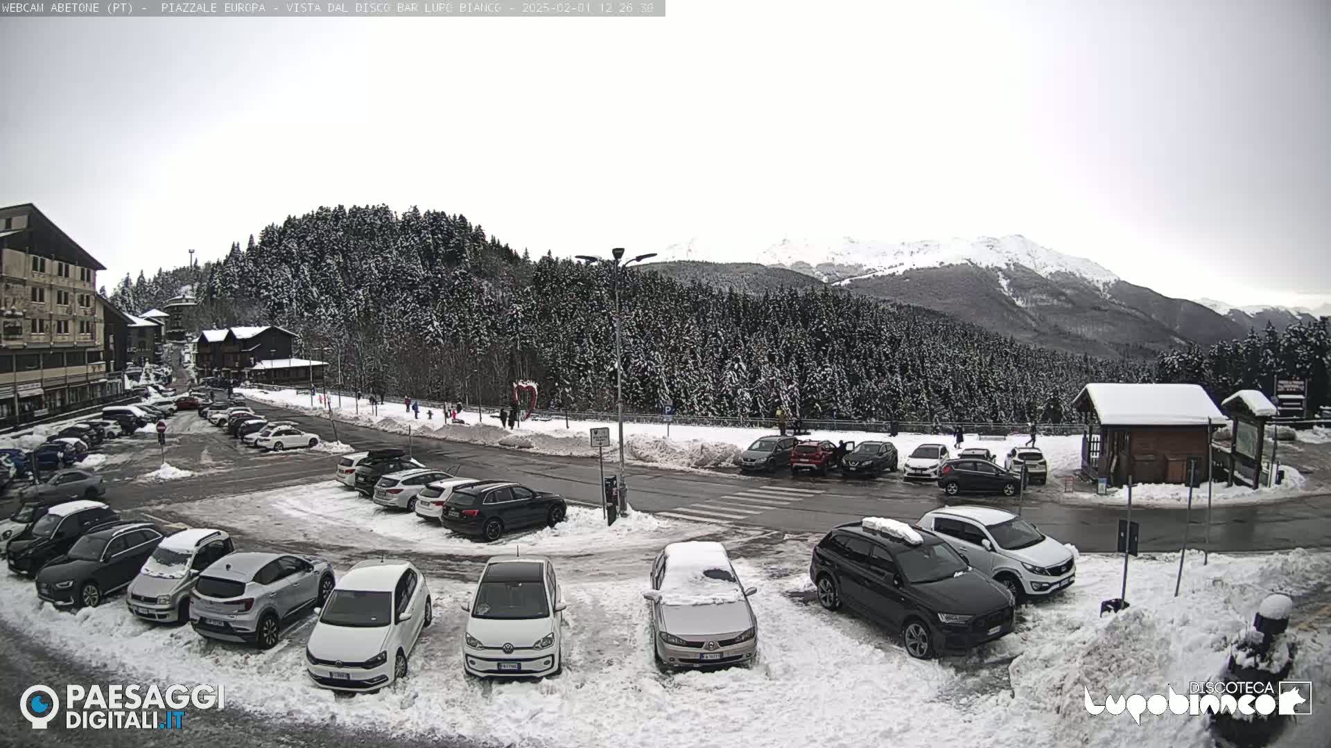 A snow-covered parking lot with cars and buildings is situated in front of a snow-dusted forest and mountains under an overcast sky.