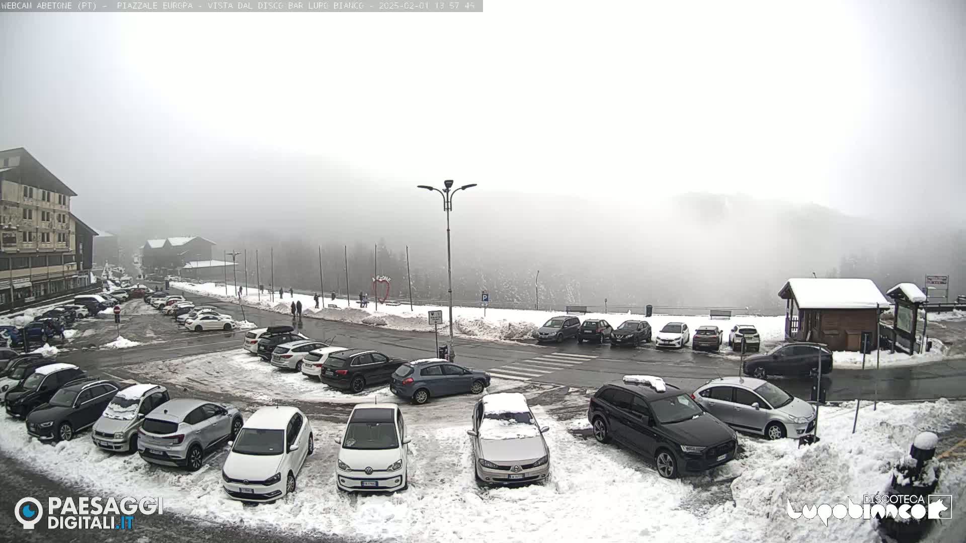 A snow-covered parking lot with numerous cars is visible in foggy conditions, with a building and a few pedestrians in the background.