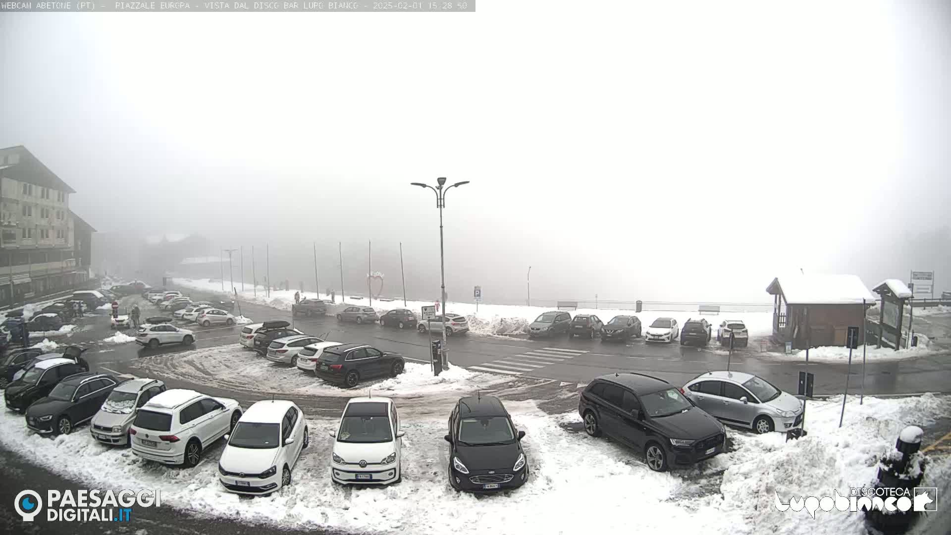 A snow-covered parking lot filled with cars is visible in a foggy mountain setting.