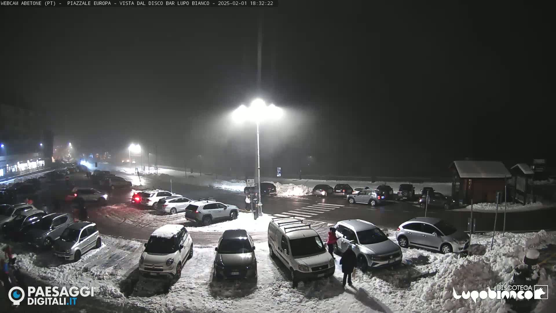 A snow-covered parking lot at night, filled with cars under dim streetlights and a light snowfall.