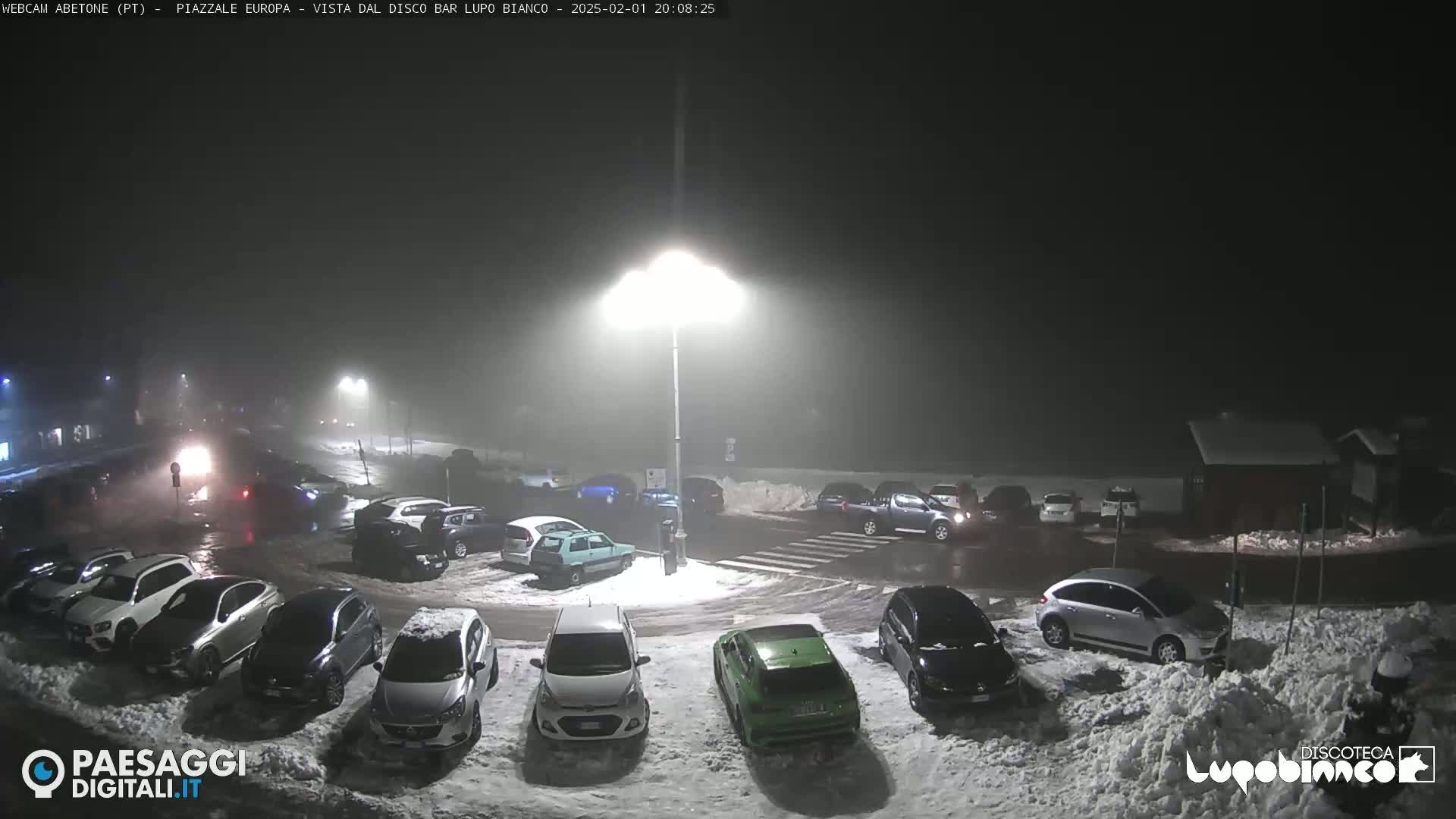 A nighttime parking lot covered in snow, illuminated by streetlights, with several cars parked and a light fog or mist in the air.