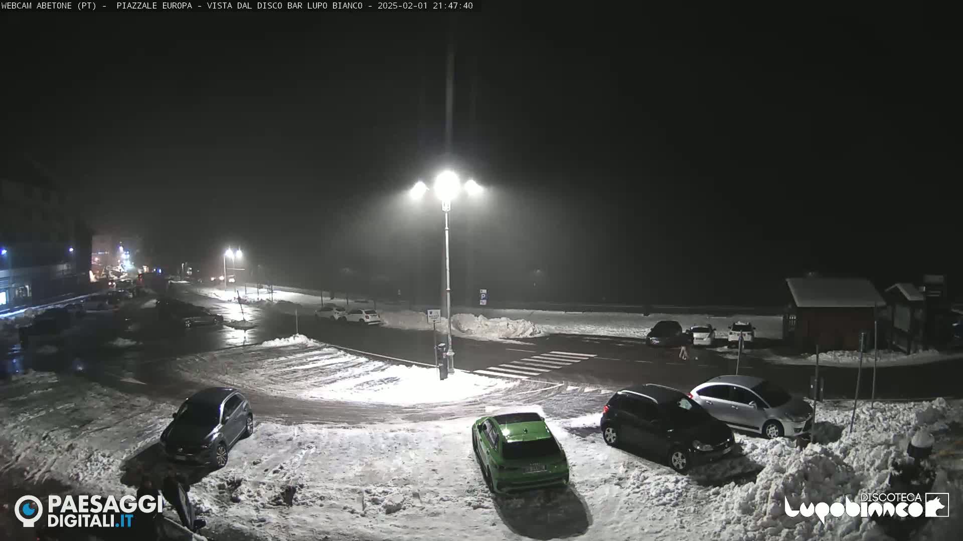 A snow-covered parking lot at night, illuminated by streetlights, with several cars parked and a few people visible.