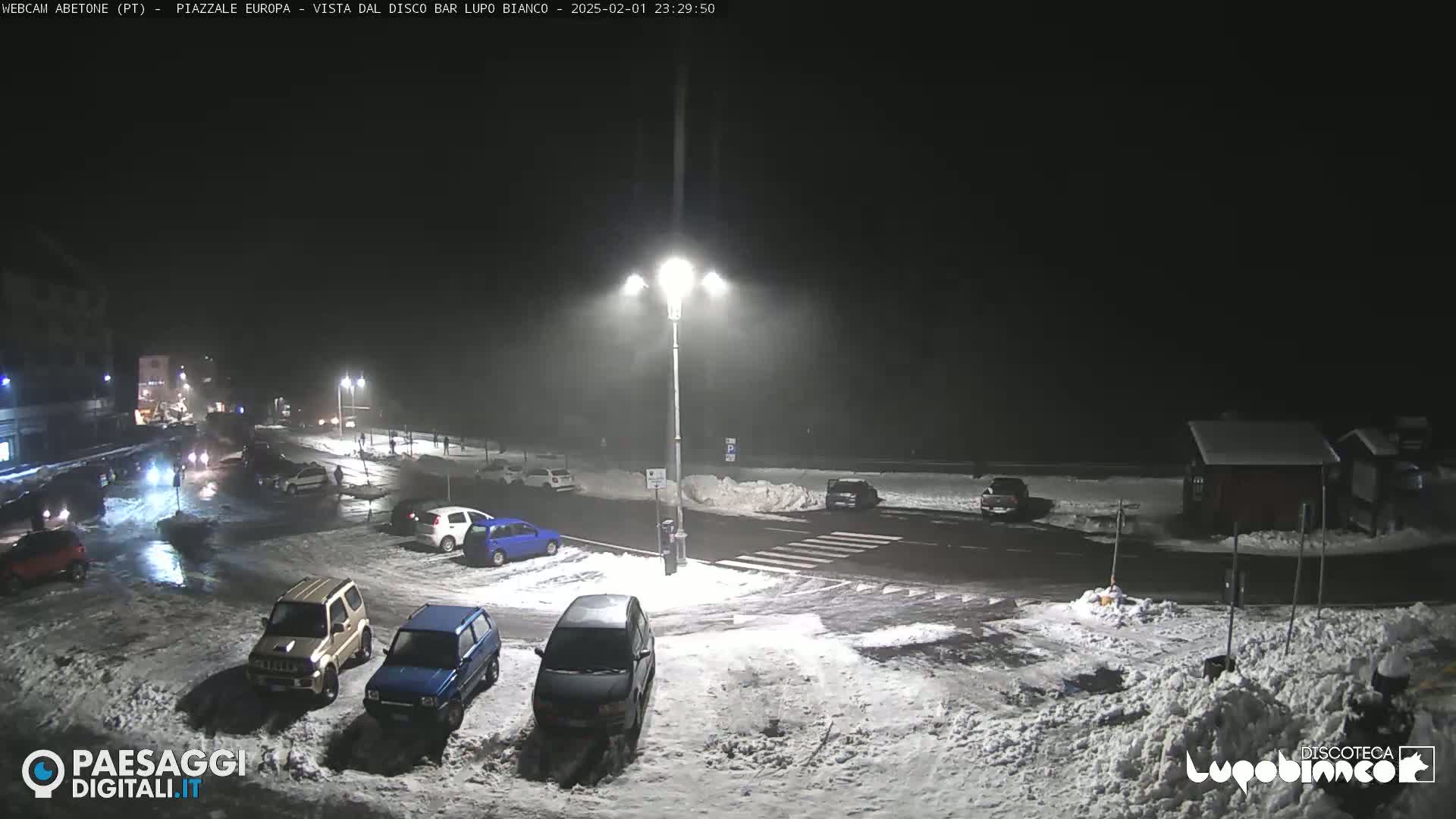 A snow-covered nighttime parking lot with several cars, illuminated by streetlights.