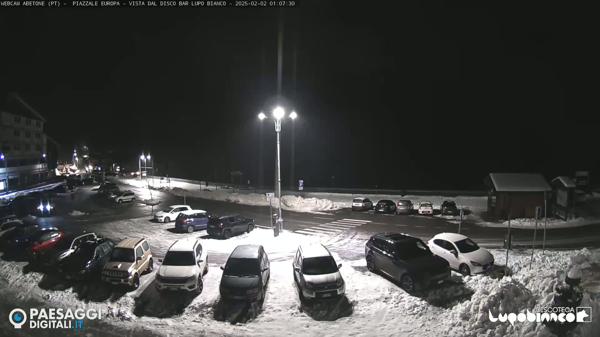 A snow-covered parking lot at night, illuminated by streetlights, with several cars parked and a building in the background.