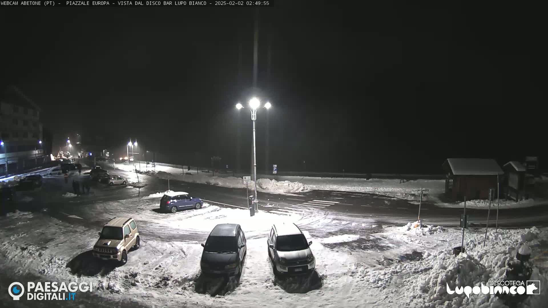 A snow-covered nighttime parking lot with several cars parked, illuminated by streetlights.