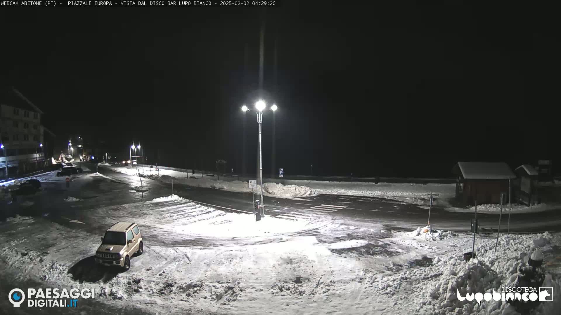 A snow-covered parking lot and road are illuminated by streetlights at night.
