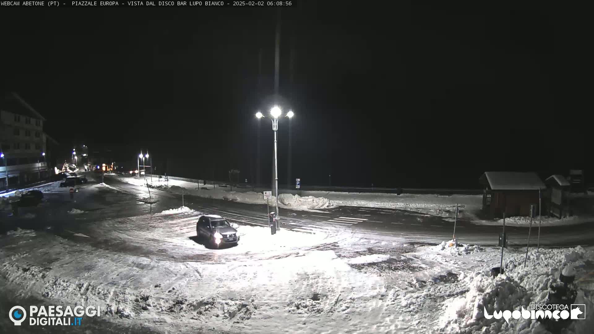 A snow-covered parking lot and road at night, illuminated by streetlights, with a vehicle parked near a corner.