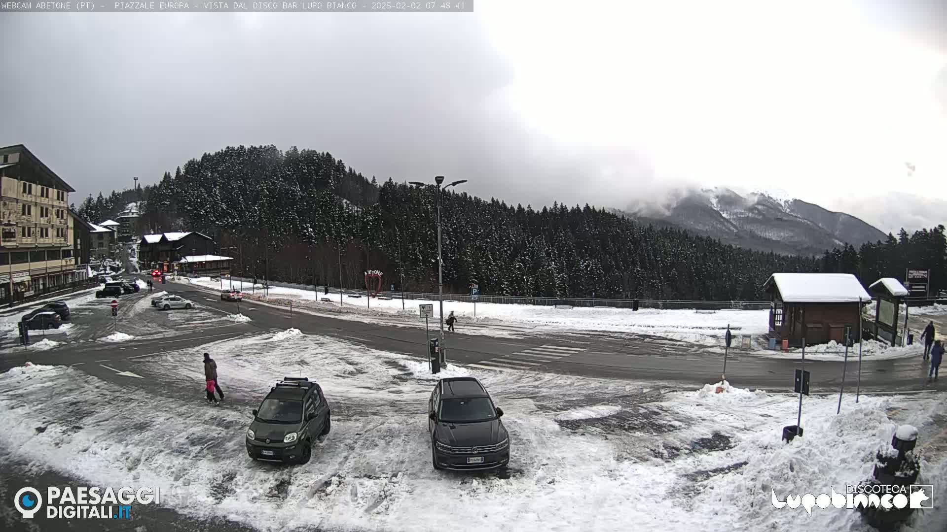 A snow-covered parking lot and roadway are situated in front of a snow-covered mountain range under an overcast sky.