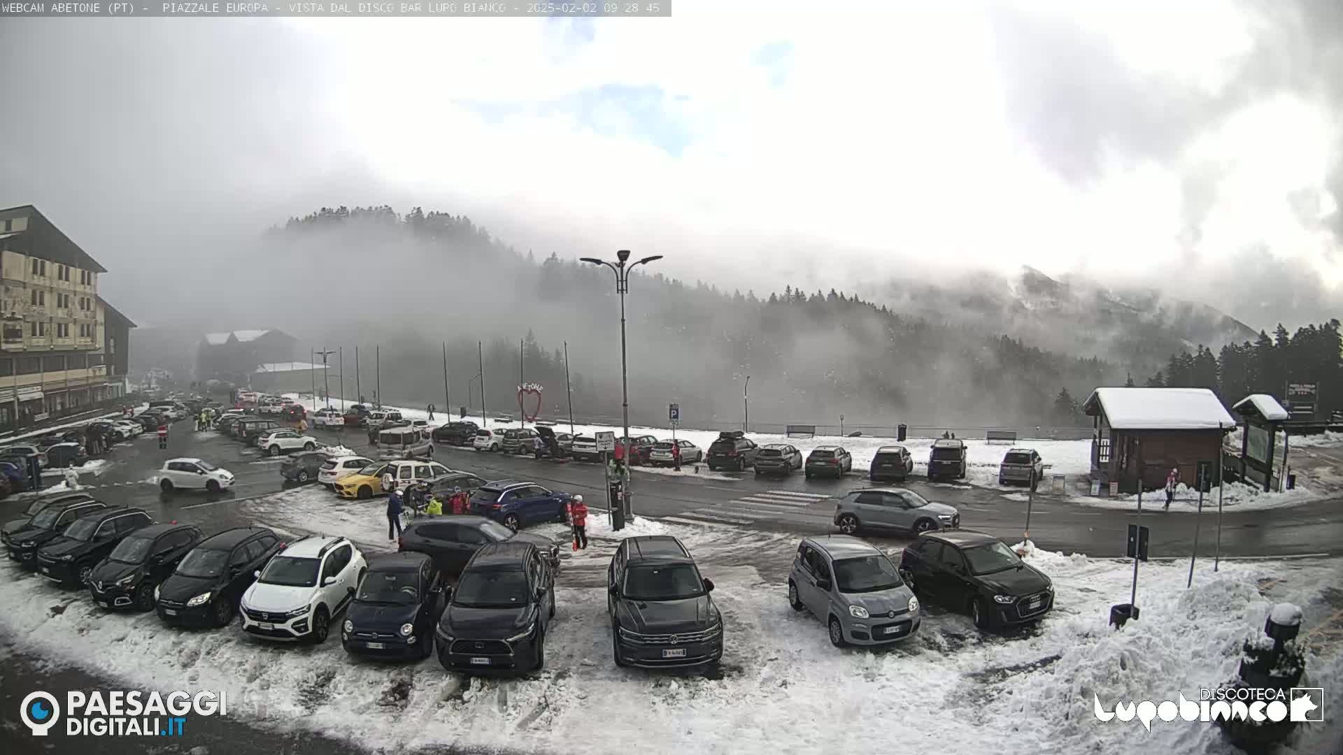 A snow-covered parking lot with many cars is situated in front of a building and a mountain shrouded in fog.