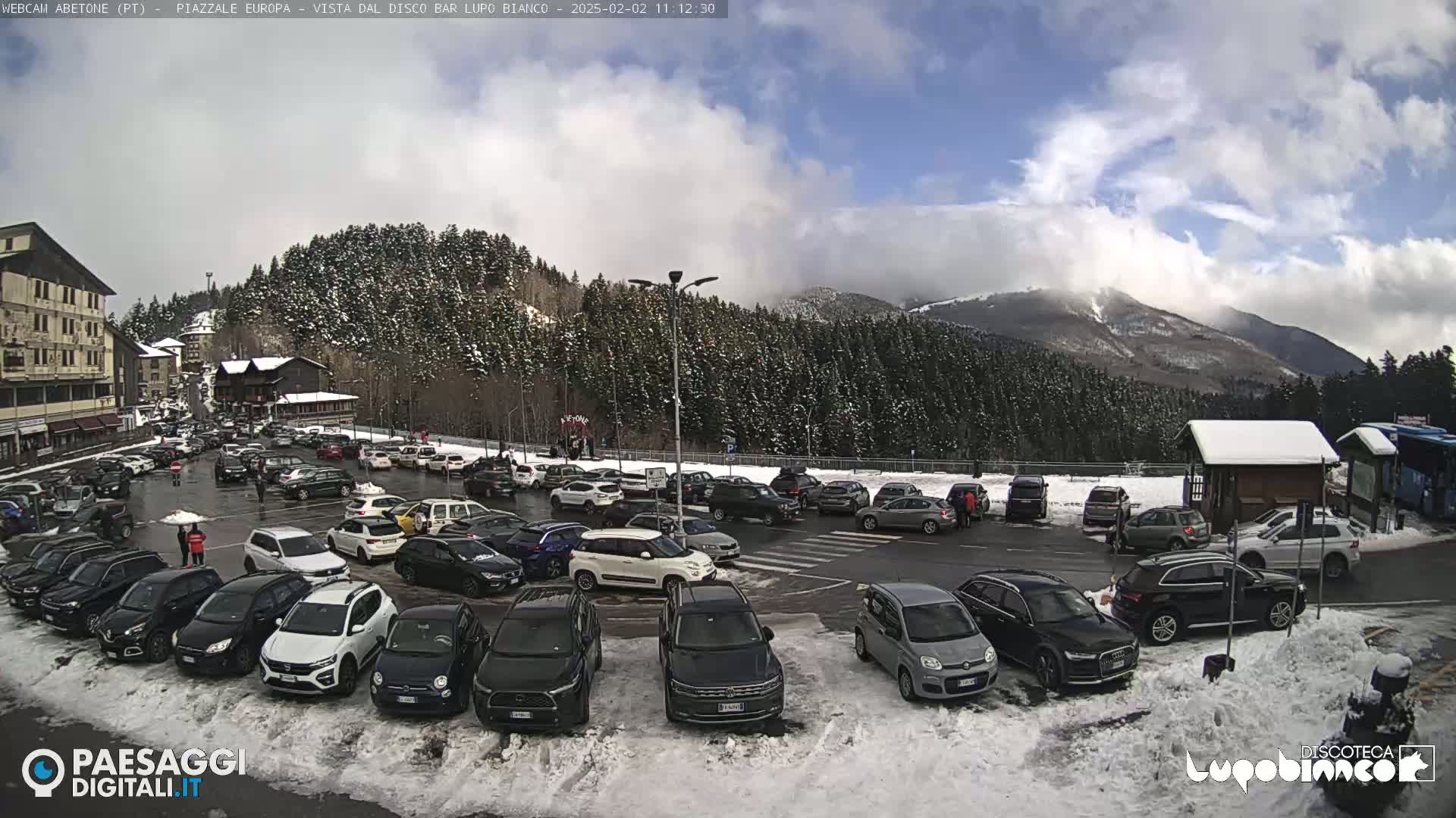 A partly cloudy day reveals a snow-covered mountain in the background, overlooking a crowded parking lot filled with cars in a snowy area.