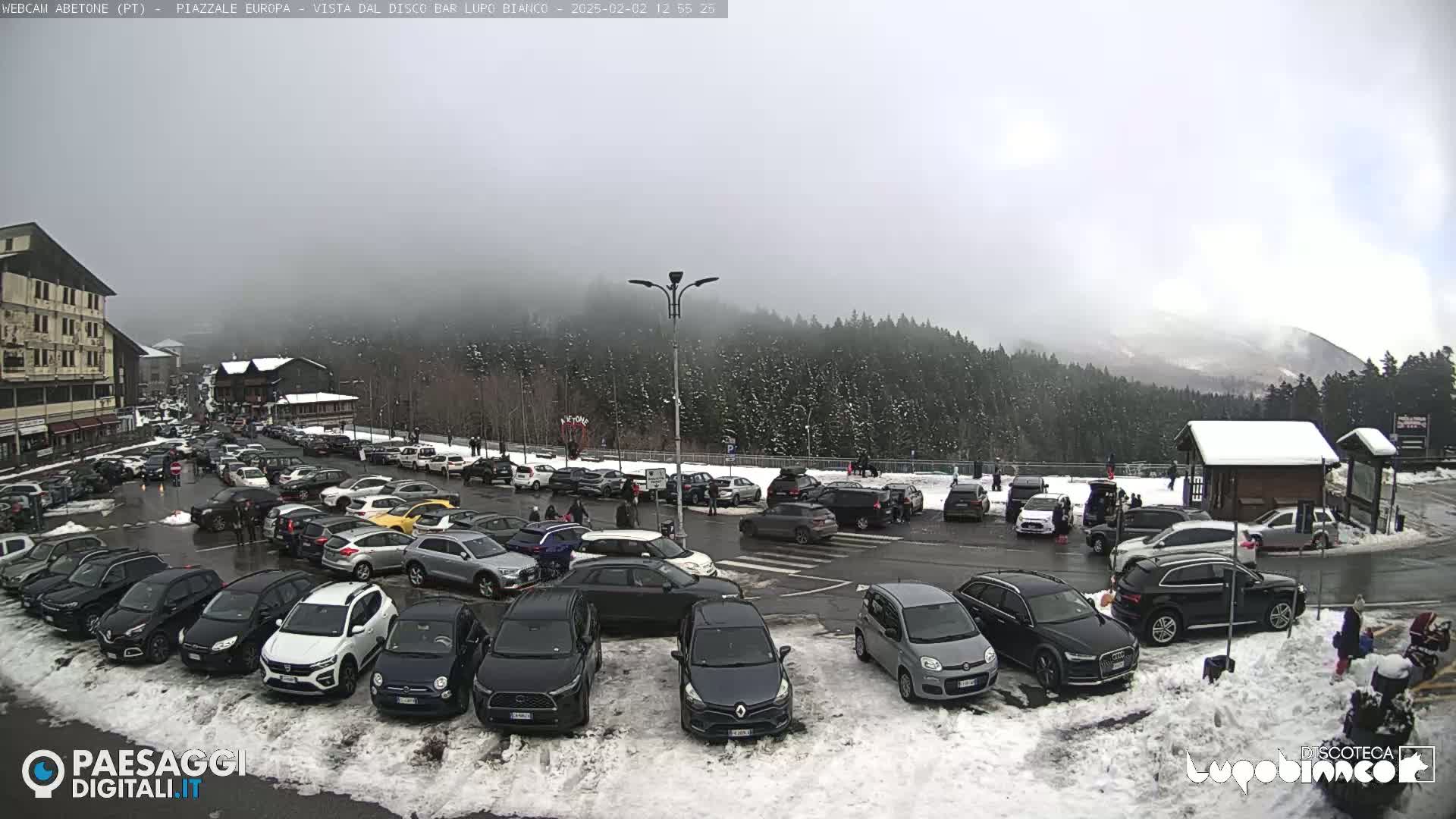 A large parking lot filled with cars is shown, covered in patches of snow, with a foggy, overcast sky and a snow-covered mountain in the background.