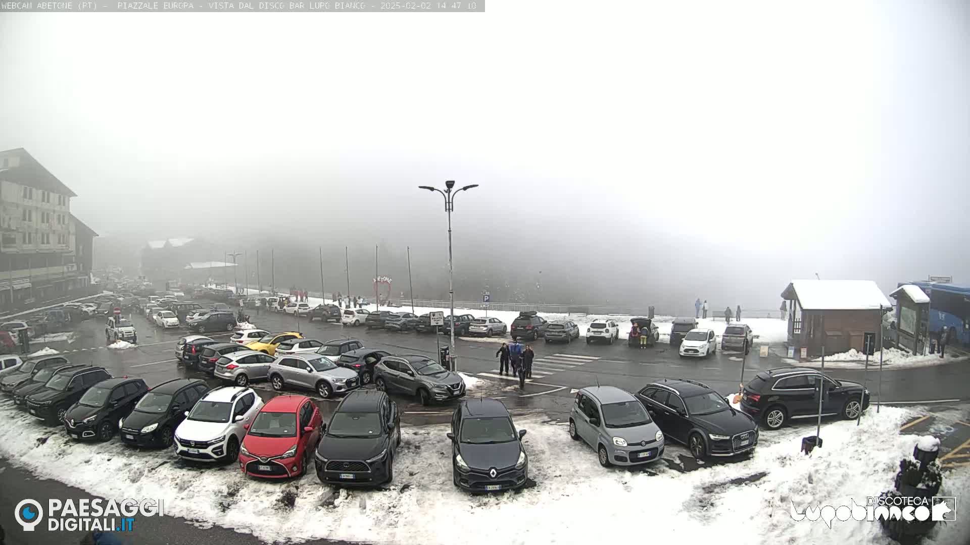 A foggy parking lot filled with cars and a few pedestrians, with patches of snow on the ground.