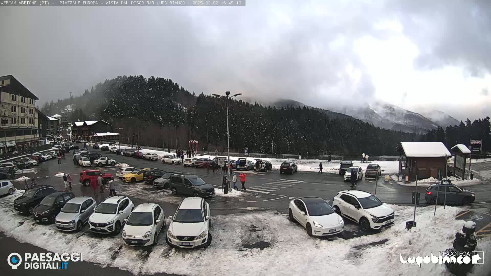 A parking lot filled with cars is situated in front of a snow-dusted mountain range under an overcast sky.