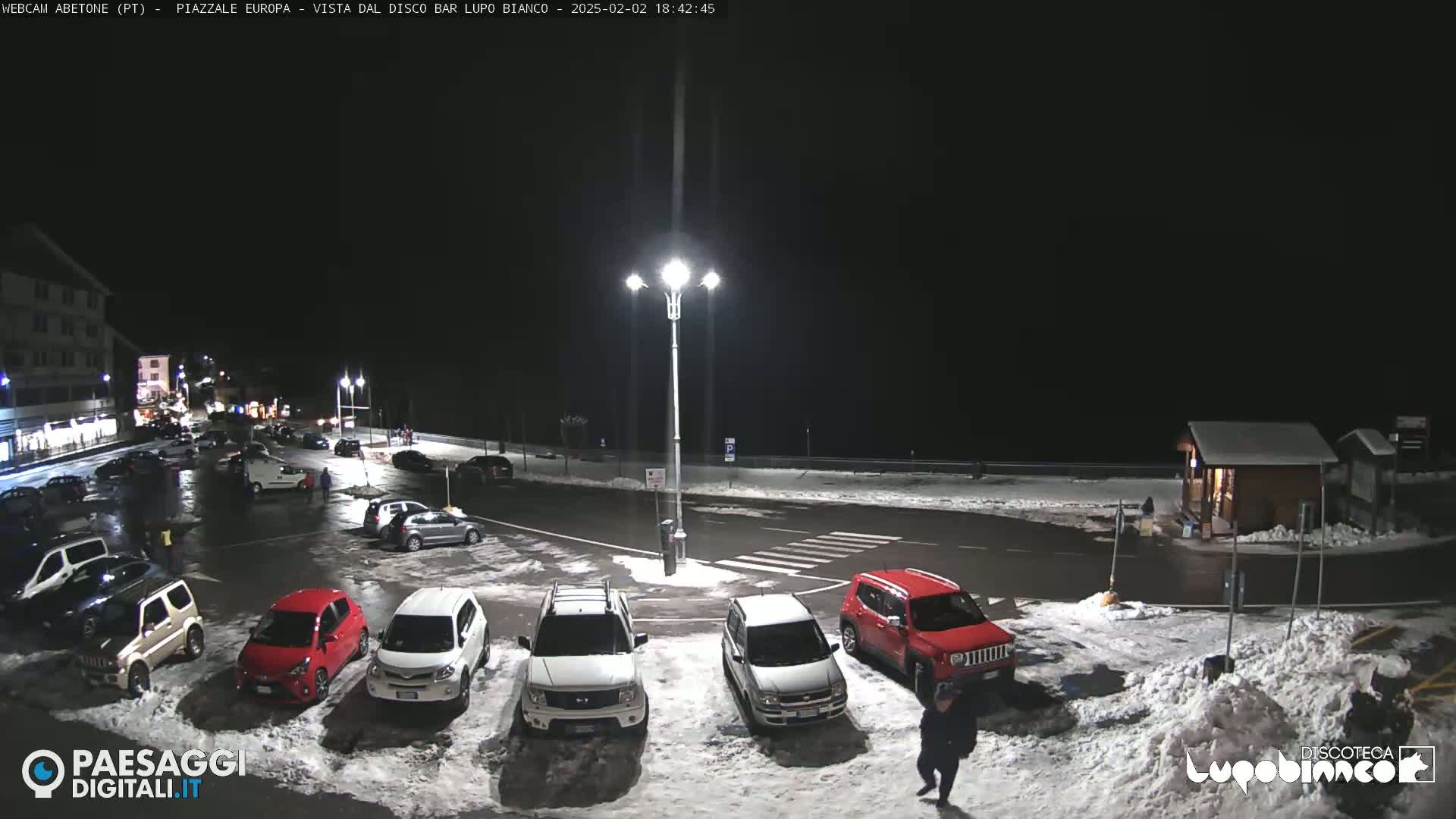 A nighttime parking lot with several cars parked in the snow, illuminated by streetlights.
