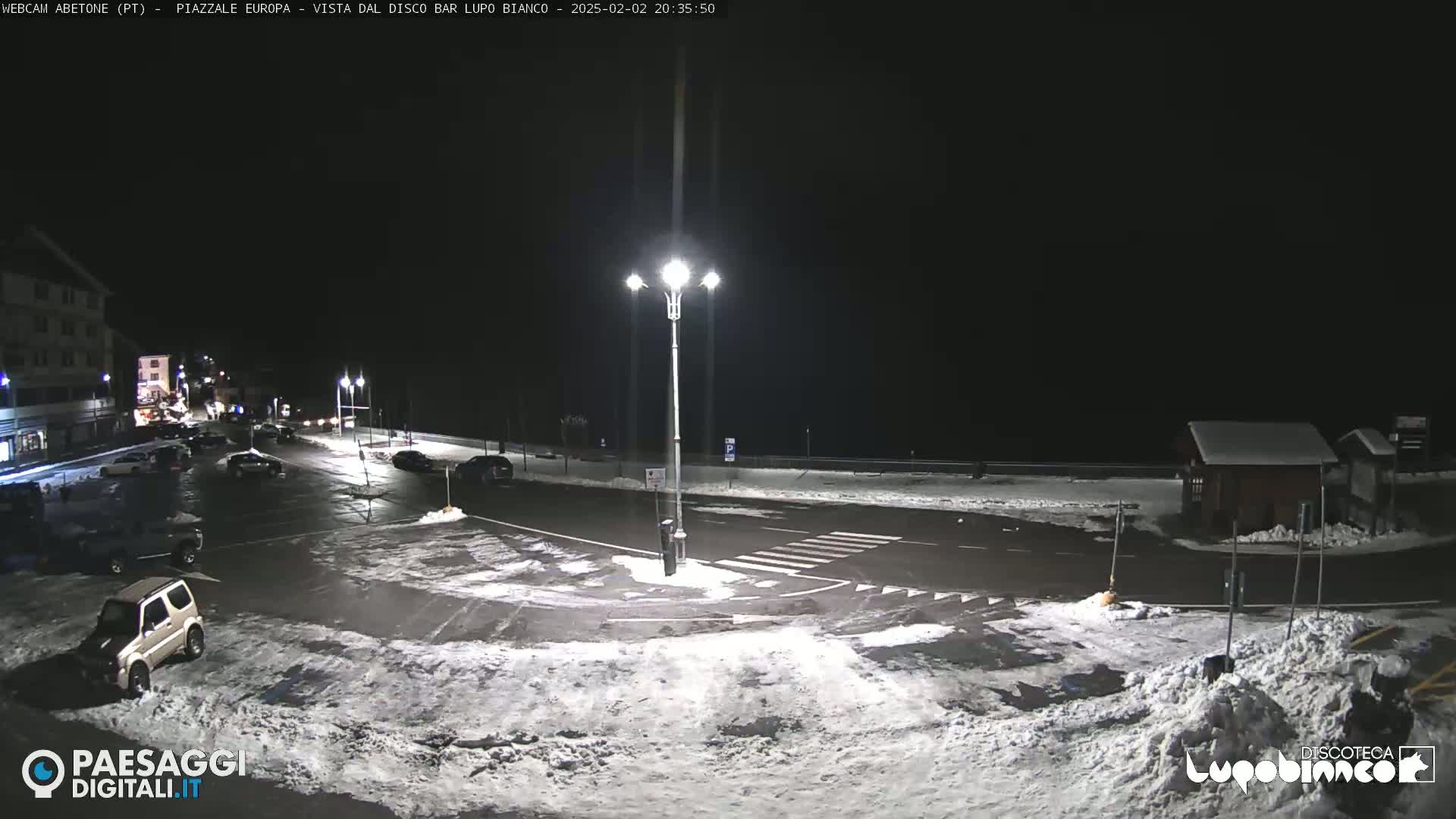 A snow-covered parking lot and roadway are illuminated at night by streetlights, with several cars parked and a building visible in the background.
