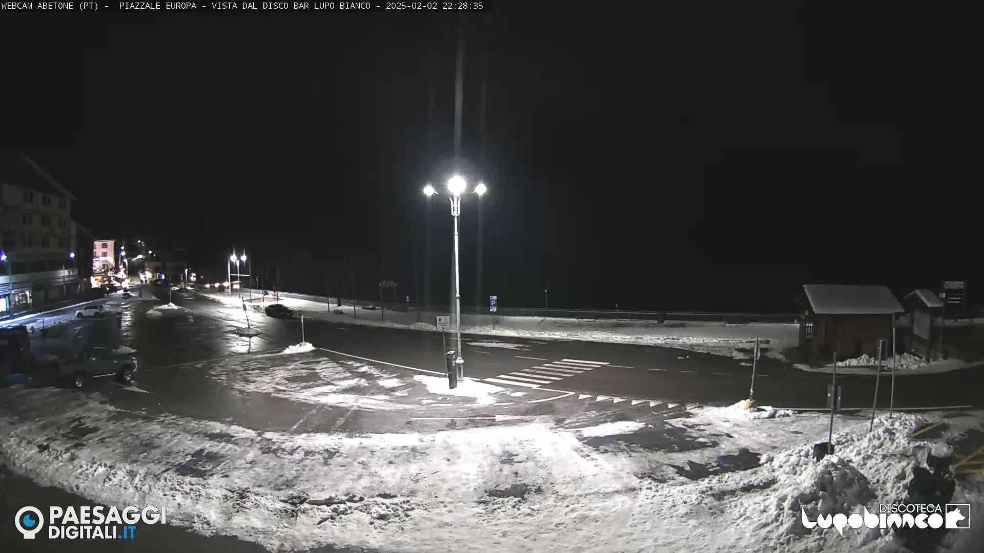 A snow-covered nighttime parking lot and road intersection are illuminated by streetlights, with a few vehicles visible and buildings in the background.