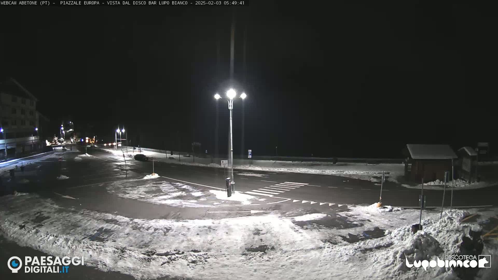 A snow-covered nighttime parking lot and roadway are illuminated by streetlights, with a building visible in the background.