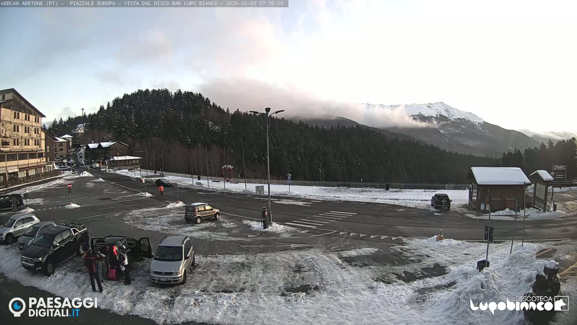 A snow-covered parking lot with several cars, surrounded by buildings and evergreen trees, with a mountain range partially obscured by clouds in the background.