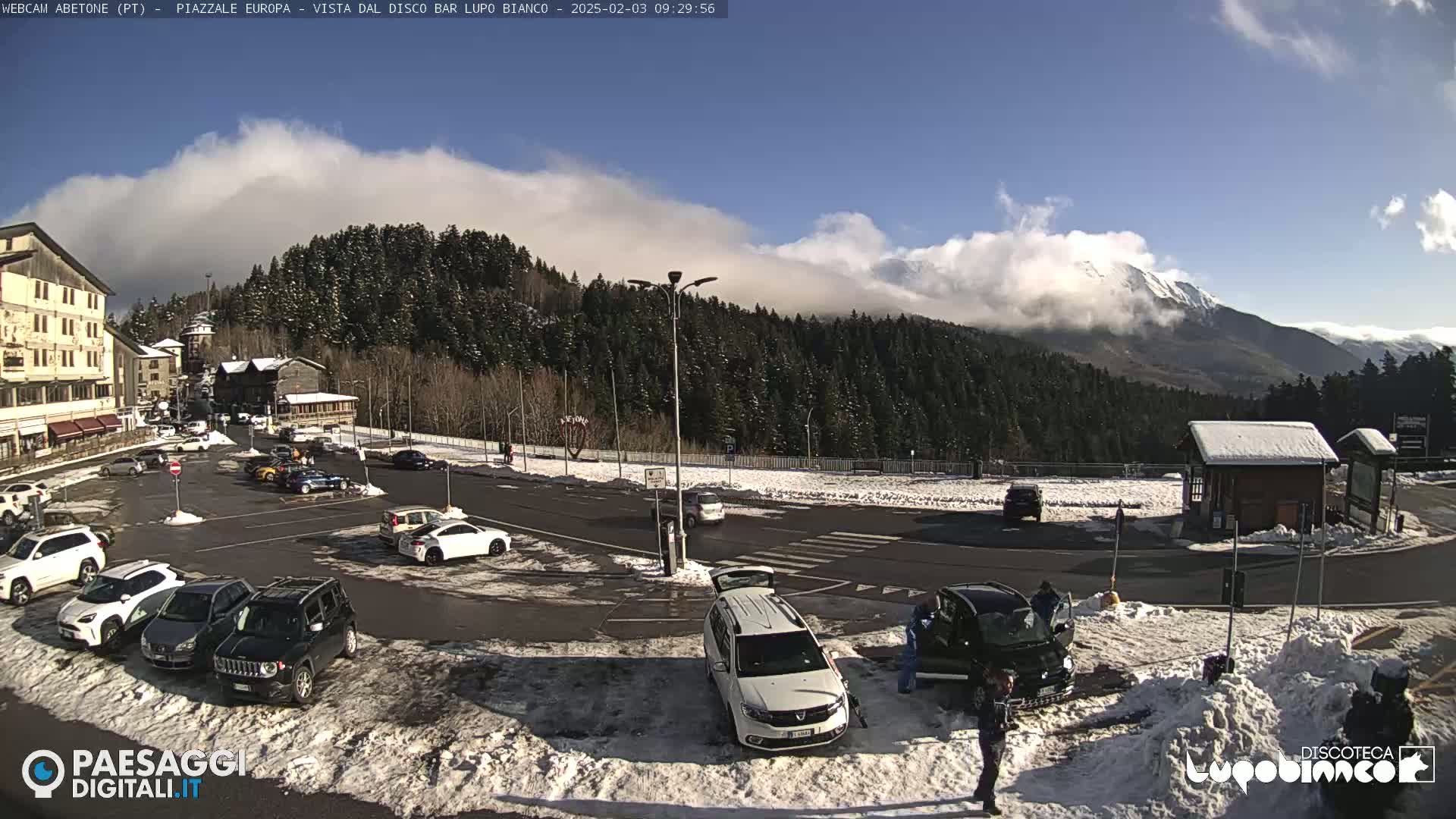 A snow-covered parking lot with several cars is situated in front of a mountain range partially obscured by clouds under a mostly sunny sky.