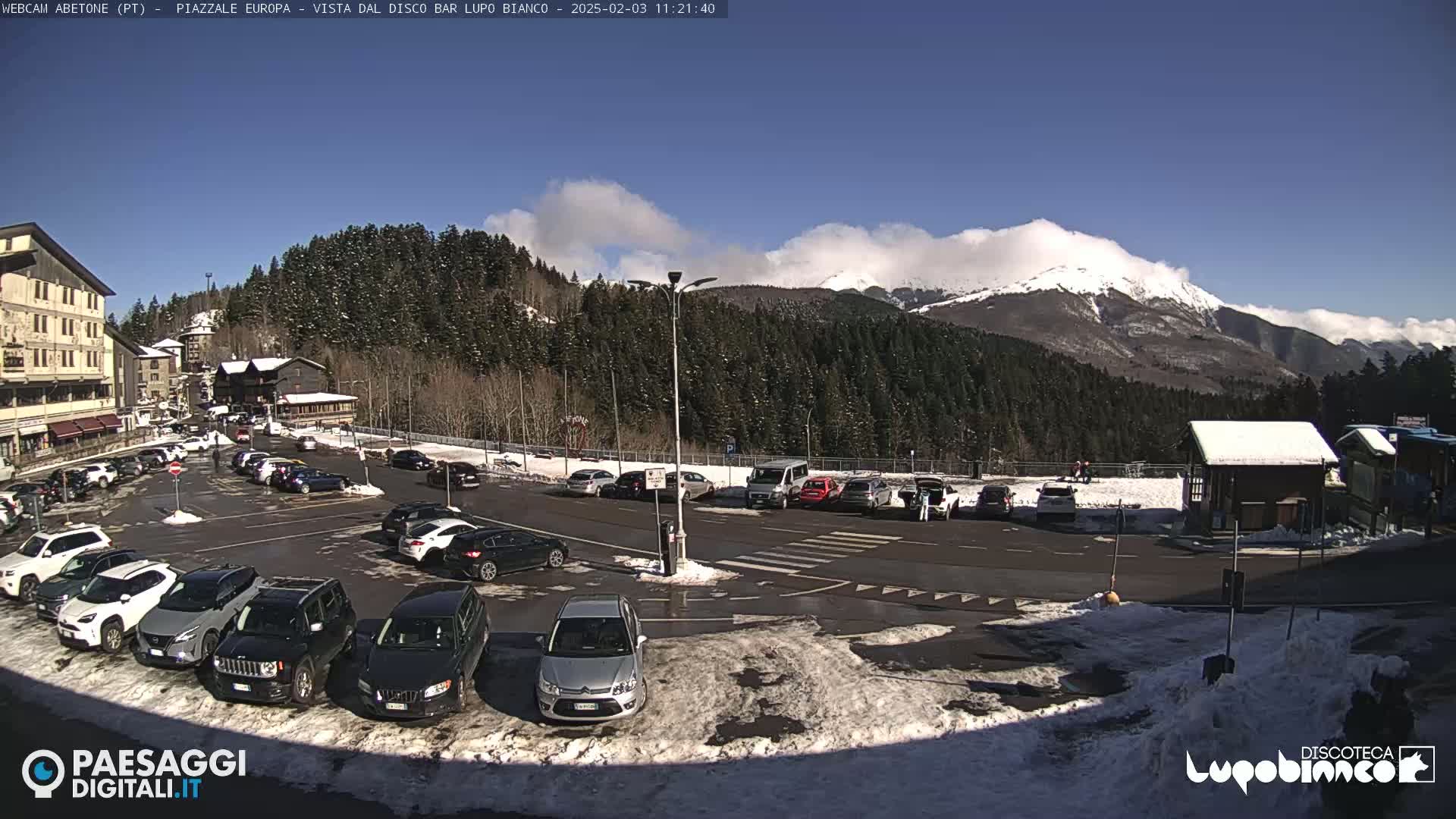 A sunny, partially cloudy day in a mountain town parking lot with snow on the ground and in the background, showing cars parked and a few people walking near buildings and snow-covered mountains.