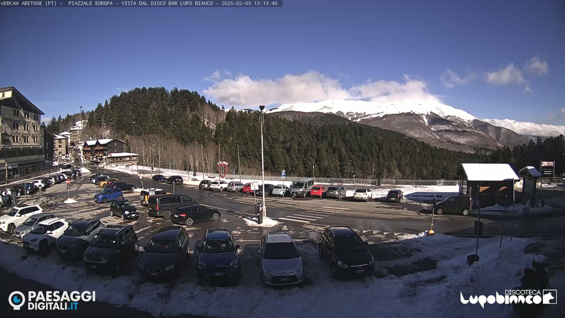 A sunny day in a snowy mountain town shows a parking lot full of cars near buildings and a snow-covered mountain in the background.