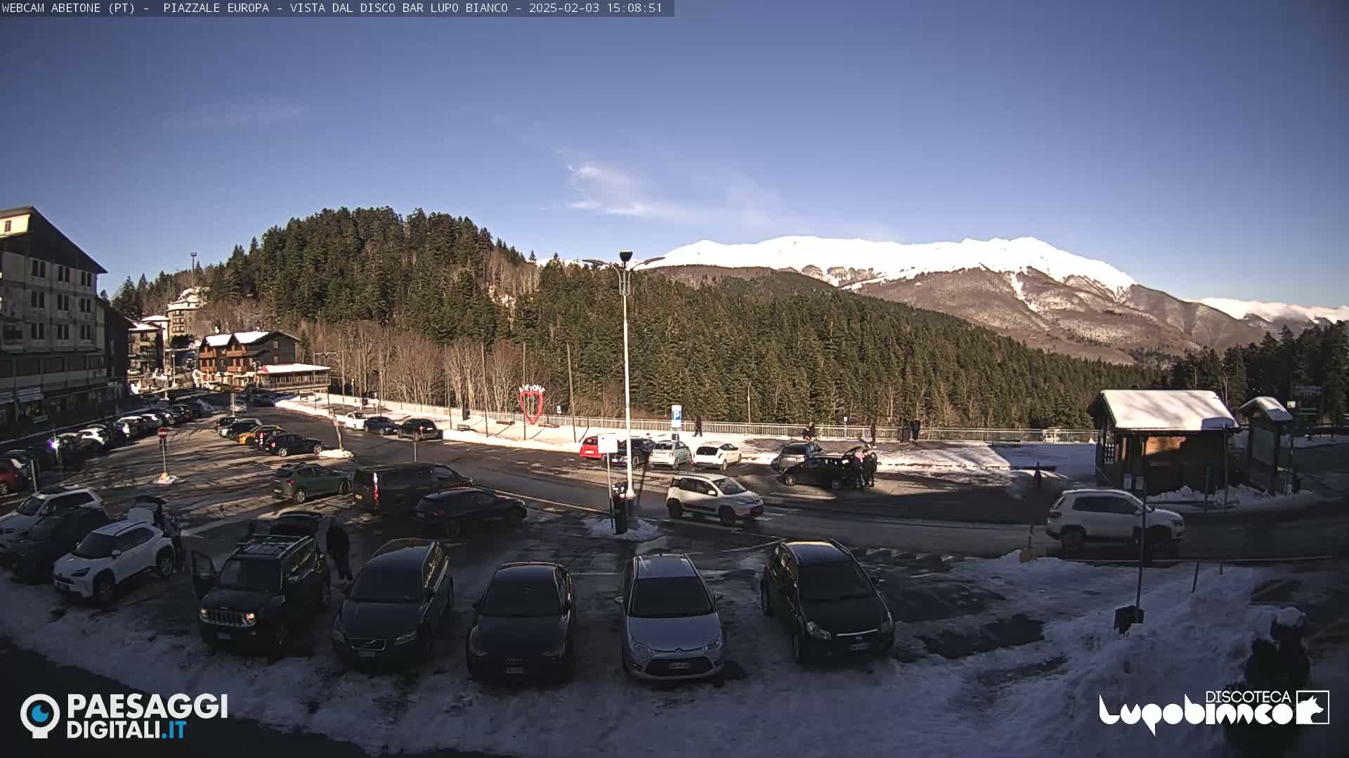 A snow-dusted parking lot filled with cars sits before a snow-capped mountain range under a clear blue sky.