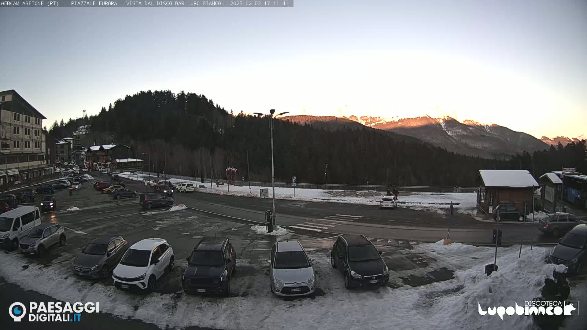 A parking lot with cars is situated in front of a building, overlooking snow-covered ground and mountains bathed in the soft light of sunset.