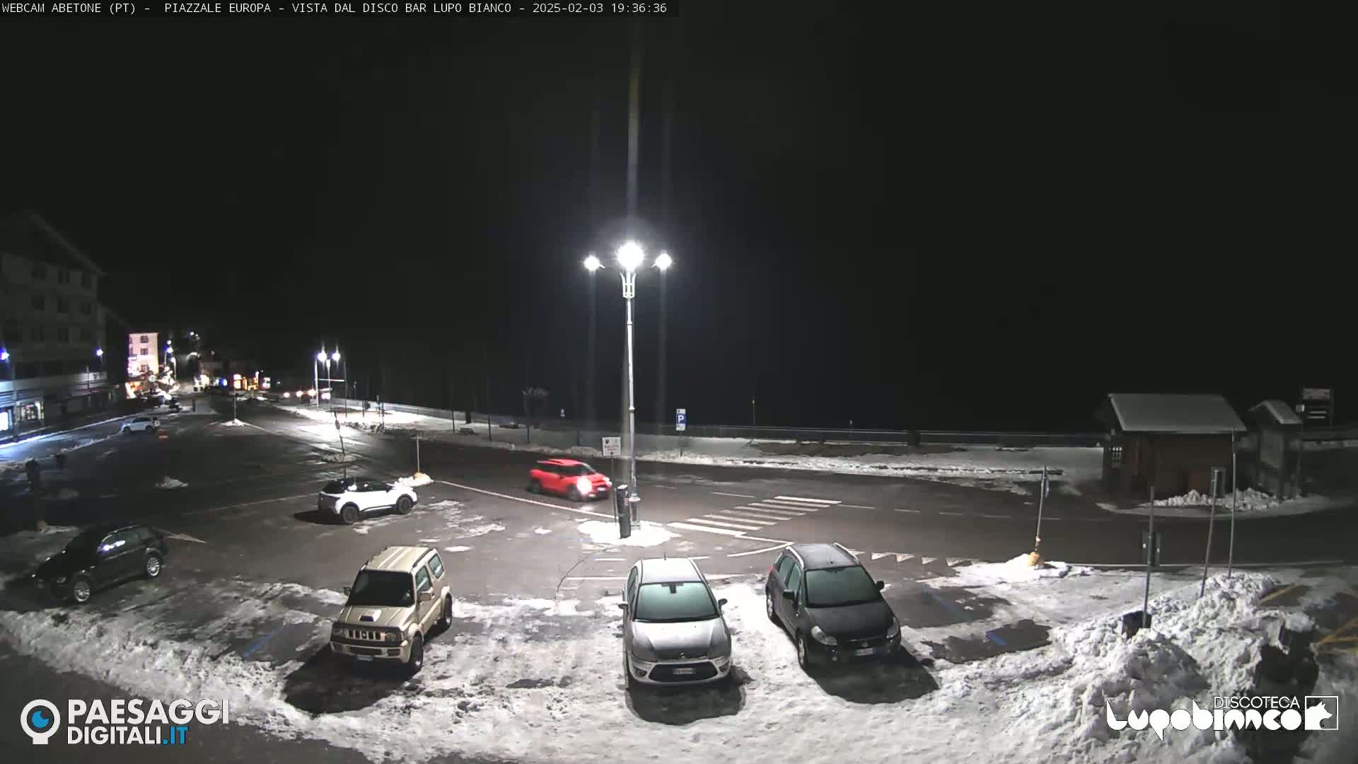 A nighttime view of a snow-dusted parking lot with several cars, under bright streetlights.