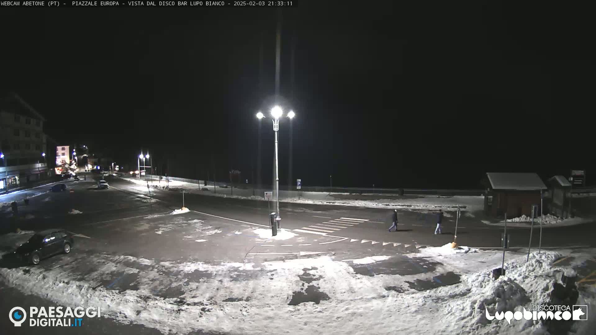 A nighttime view of a snow-covered parking lot and roadway, illuminated by streetlights, with a few people walking and some cars parked.