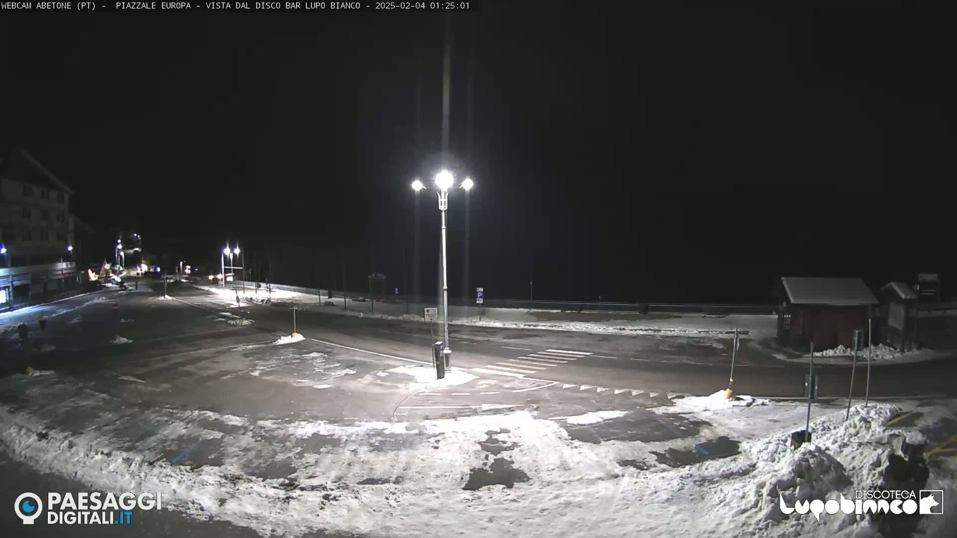 A snow-covered nighttime parking lot and roadway are illuminated by streetlights.