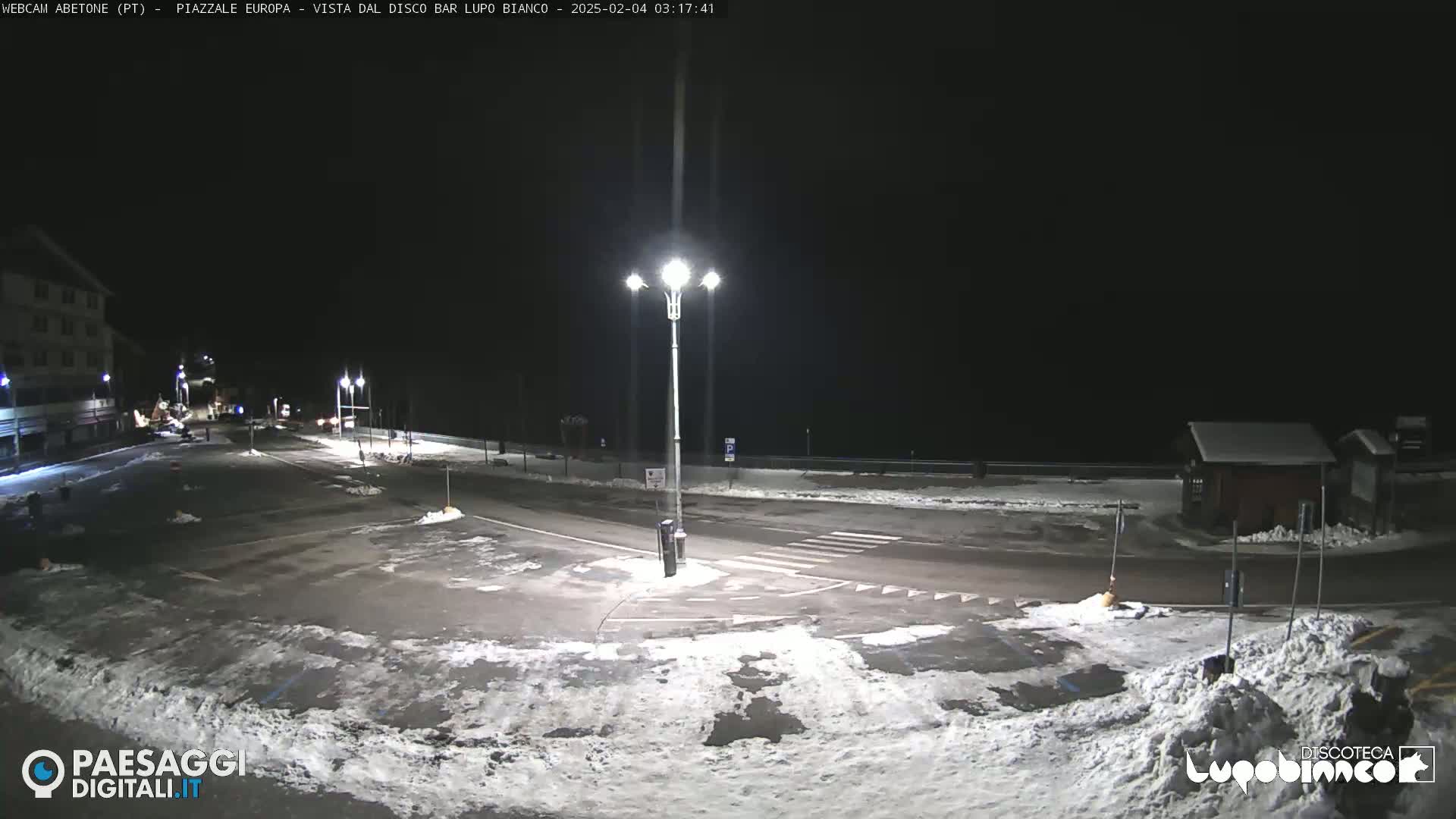 A snow-covered parking lot and road are illuminated by streetlights at night.